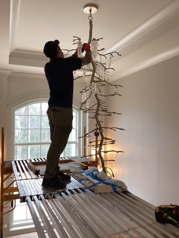 A person stands on scaffolding while installing or adjusting a large, branch-shaped chandelier hanging from the ceiling in a room with a large window.