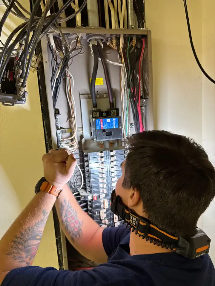 A person inspects or works on an open electrical breaker panel, wearing a headlamp and an orange wristwatch.