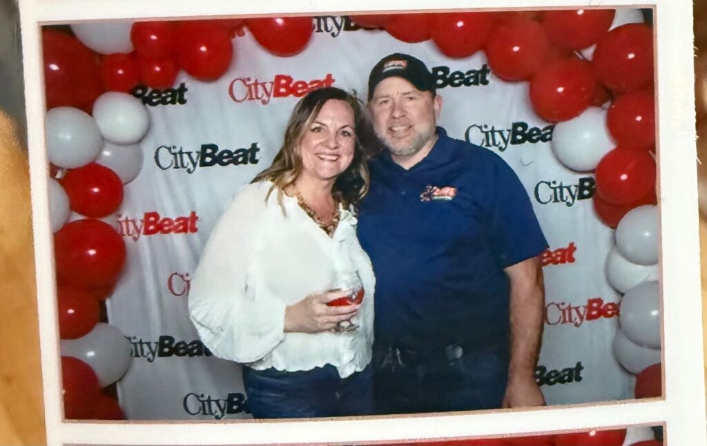 Two adults stand smiling together in front of a CityBeat backdrop framed with red and white balloons; the woman holds a drink and the man wears a cap.