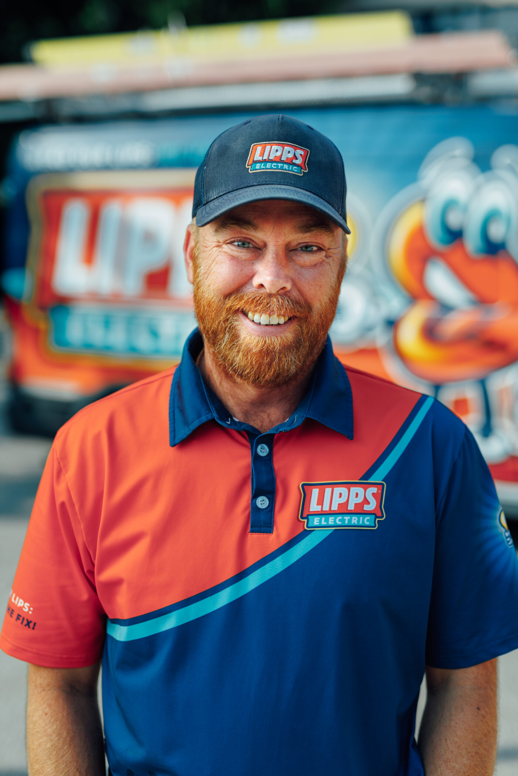 A man with a red beard in a Lipps Electric uniform and cap smiles, standing in front of a colorful Lipps Electric van with cartoon graphics.