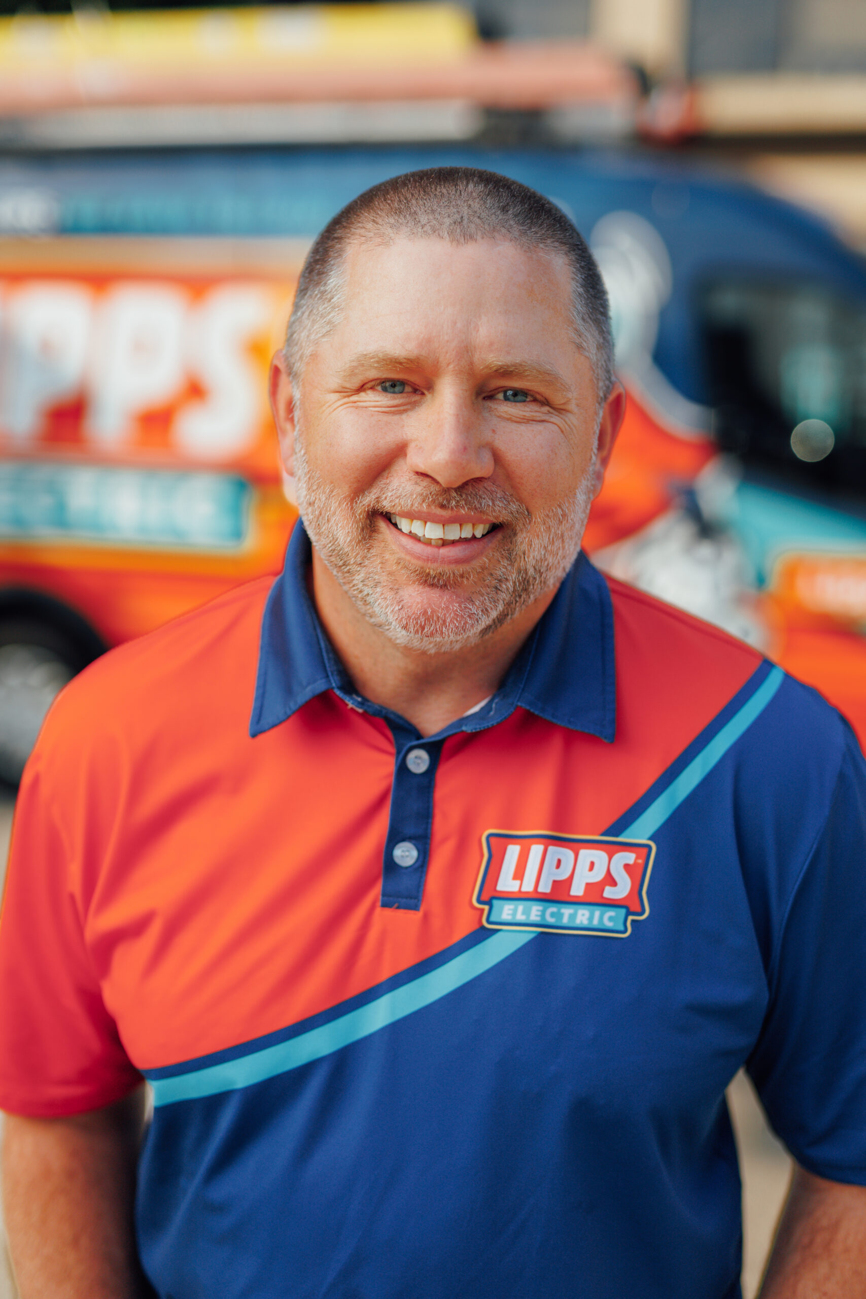Smiling man in a red and blue Lipps Electric shirt stands in front of a branded Lipps Electric service van.