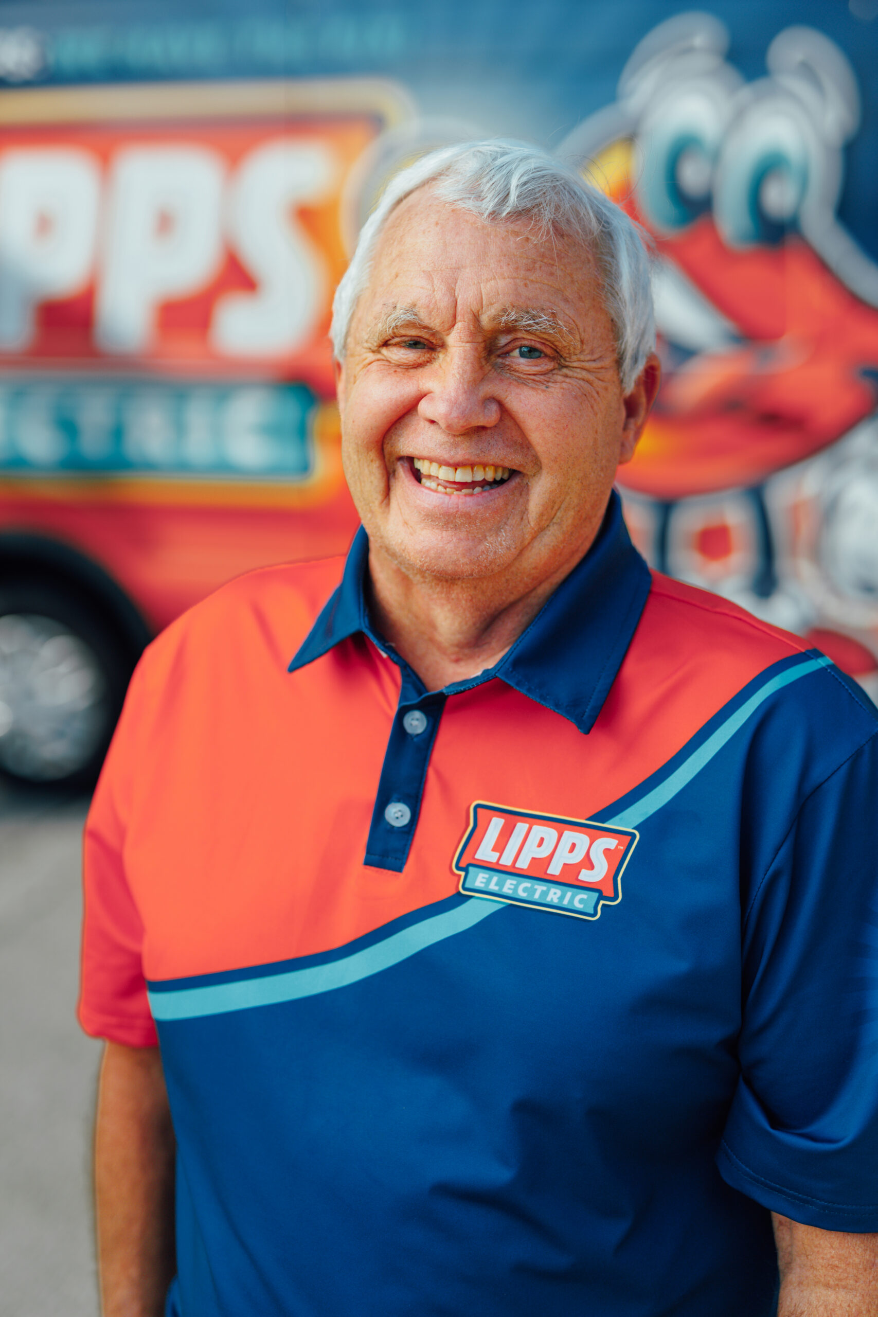 Older man with white hair smiling, wearing a Lipps Electric shirt, standing in front of a blurred Lipps Electric vehicle with colorful branding.