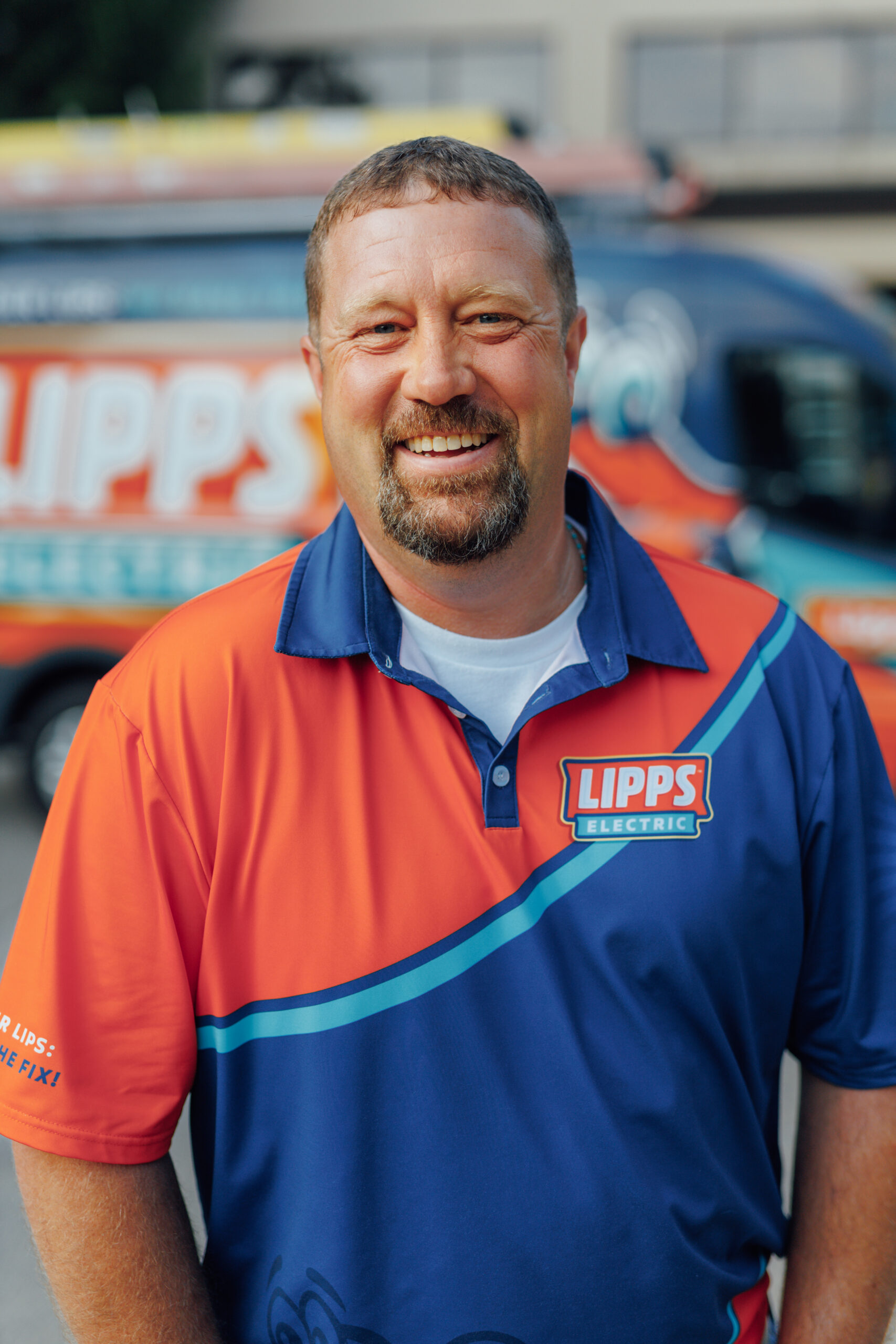 A smiling man with a goatee wearing a Lipps Electric orange and blue polo shirt stands in front of a company van.