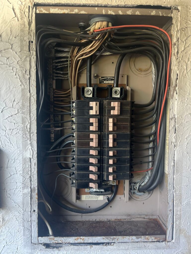 An open electrical breaker panel with multiple black breakers, wires connected at the top and sides, and a textured wall surrounding the box.