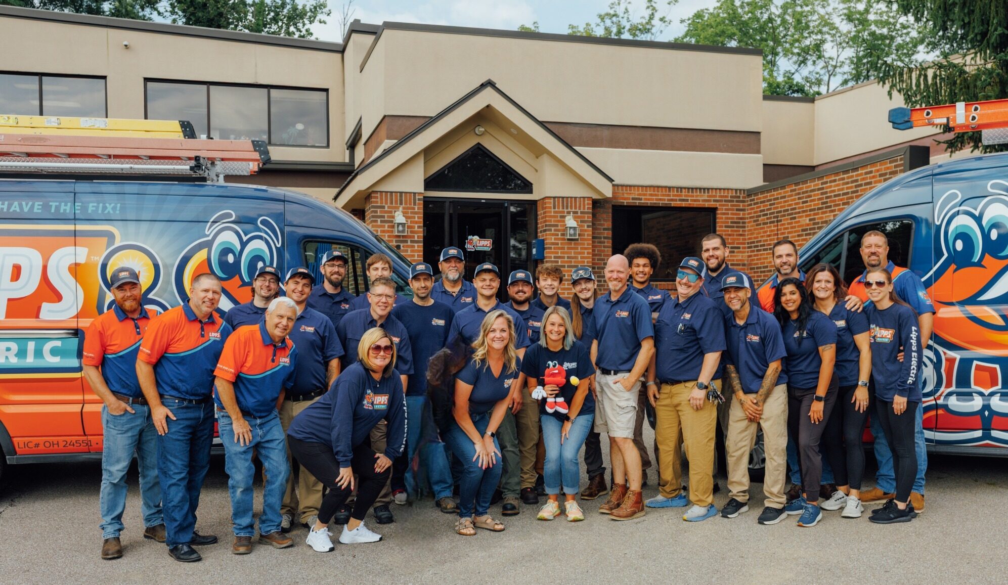 A group of people in branded shirts pose and smile outside a building between two colorful vans with cartoon mascots and electrical equipment on their roofs.