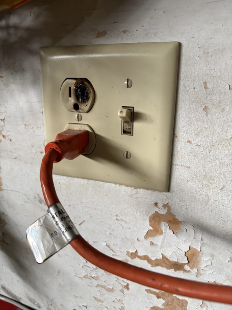 A damaged wall outlet with a burnt upper socket, a red power cord plugged into the lower socket, and a switch on a peeling, off-white wall plate.